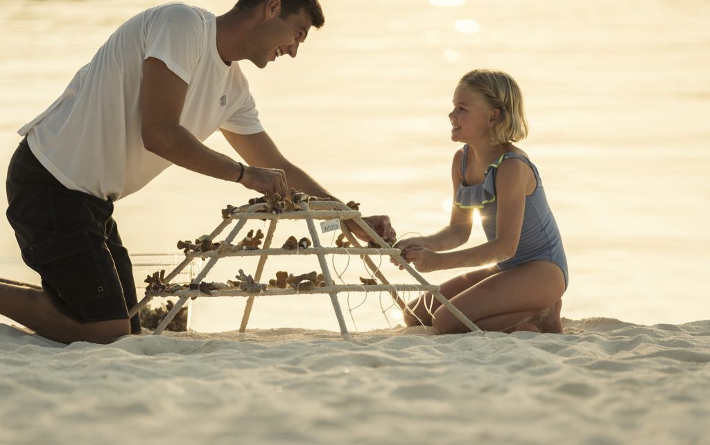Erwachsener und Kind am Strand der Malediven bauen pyramidenförmiges Metallgestell mit Sand