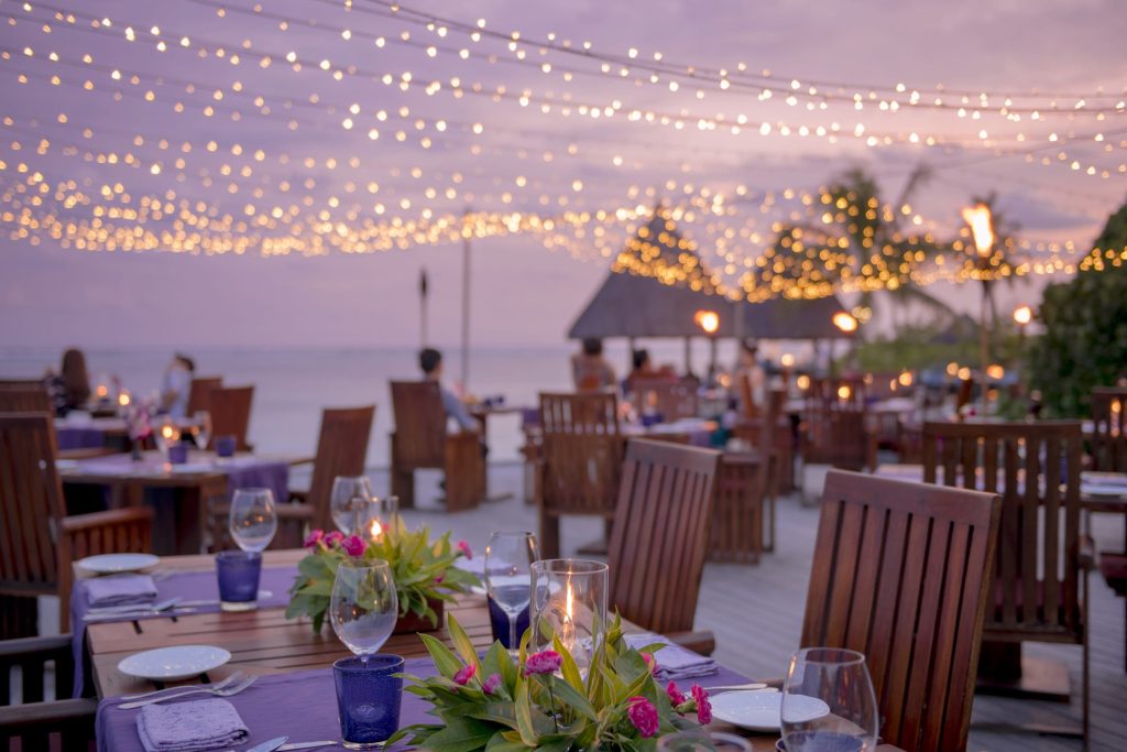 Außenbereich des Four Seasons Kuda Huraa Restaurants mit Holztischen, violetten Tischdecken und Lichterketten am Abend