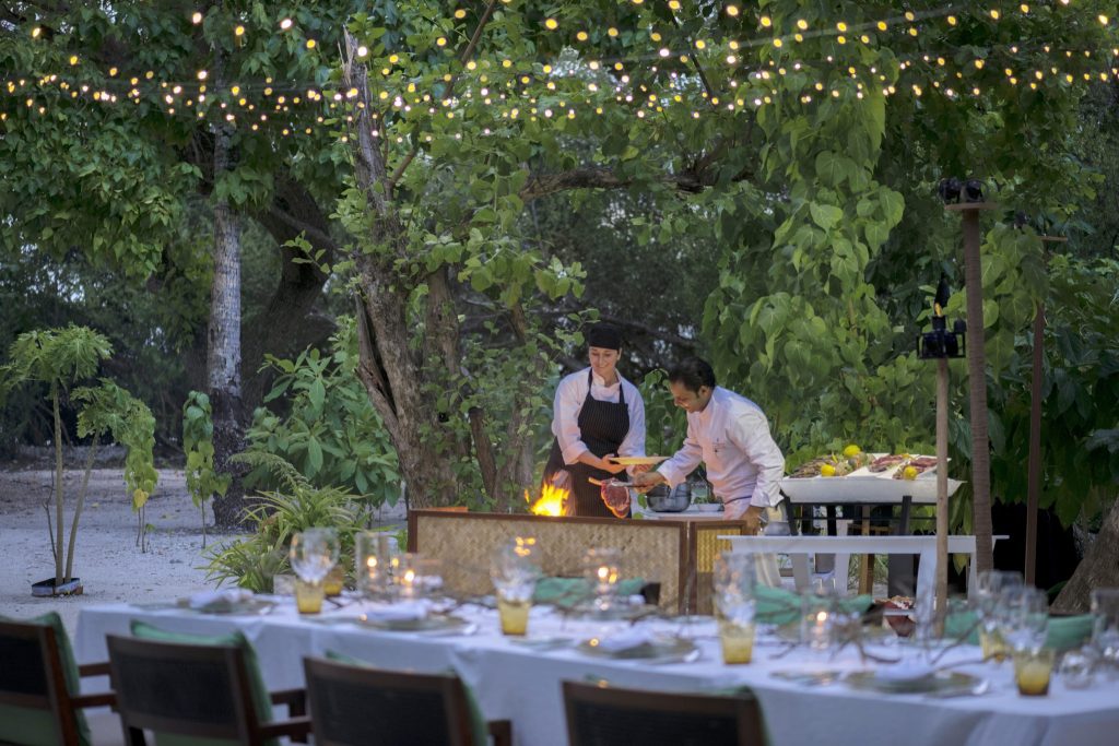 Zwei Köche in Kochkleidung am Grilltisch unter Blätterdach mit Lichterkette und gedeckten Tischen