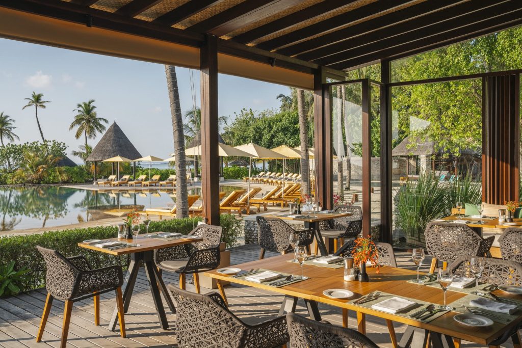 Offener Restaurantbereich mit Holztischen und geflochtenen Stühlen, Pool und tropischer Landschaft im Hintergrund