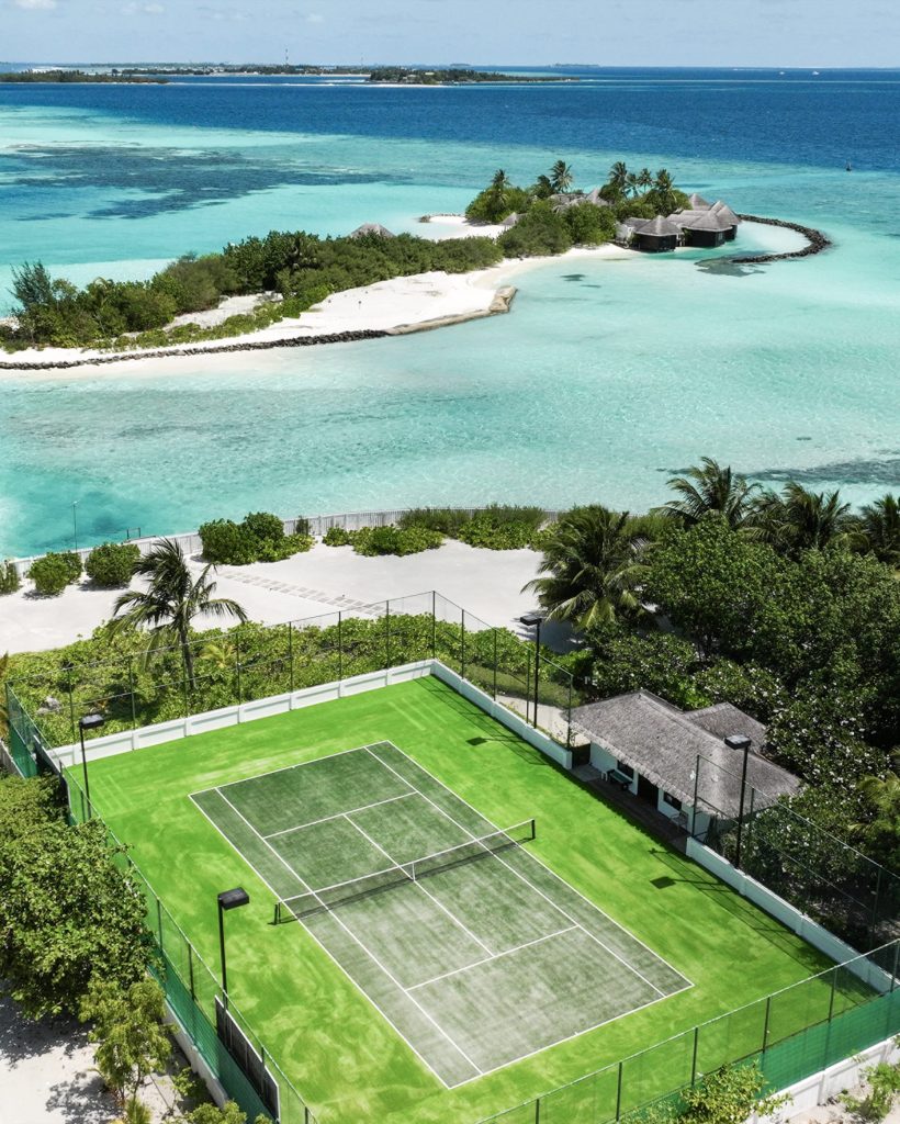 Tennisplatz mit grünem Kunstrasen, Netz und Zaun, umgeben von Palmen und tropischer Vegetation mit Meer und Insel.