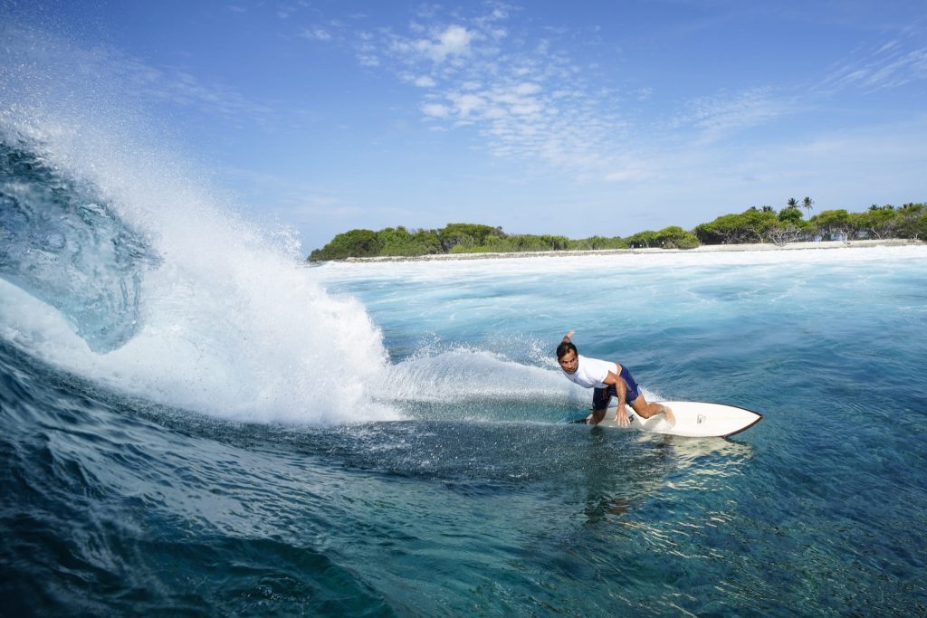 Surfer gleitet über eine Welle im blauen Ozean vor grüner Insel im Four Seasons Resort Maldives at Kuda Huraa