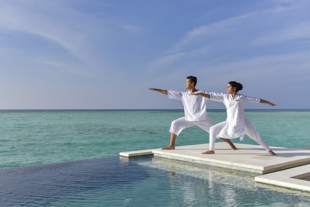 Zwei Personen in weißen Outfits üben Yoga auf weißem Steg neben Infinity-Pool mit Meer im Hintergrund