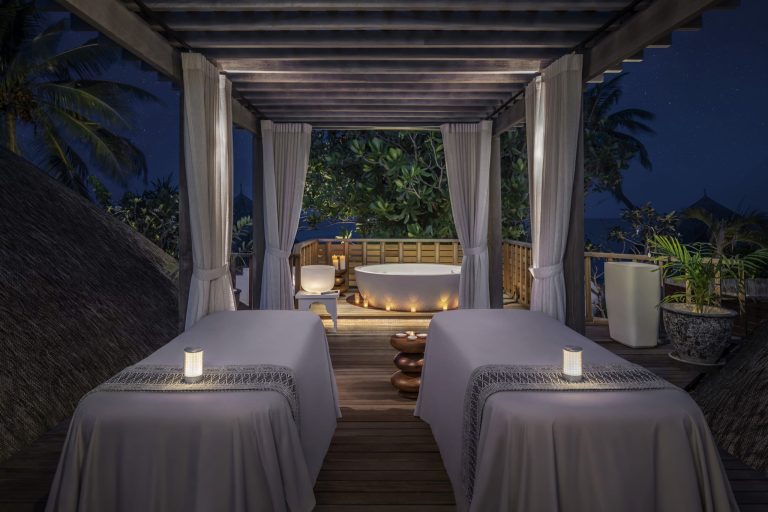 Offener Spa-Bereich mit zwei Massageliegen, runder Badewanne, Holzverkleidung und dichter Vegetation bei Nacht