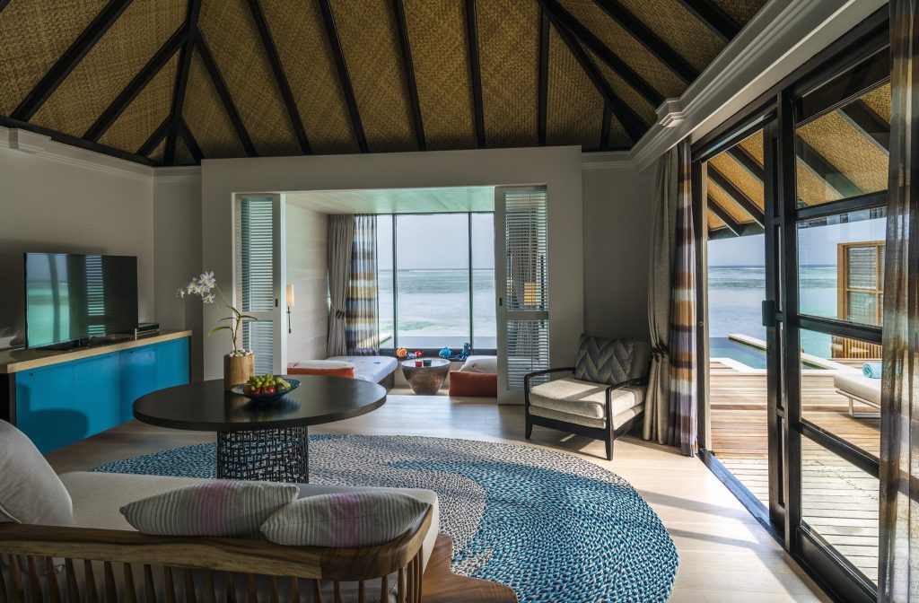 Hotelzimmer mit großem Glasfenster, blau-weißem Teppich, Holzmöbeln und Blick auf Terrasse mit Pool und Meer