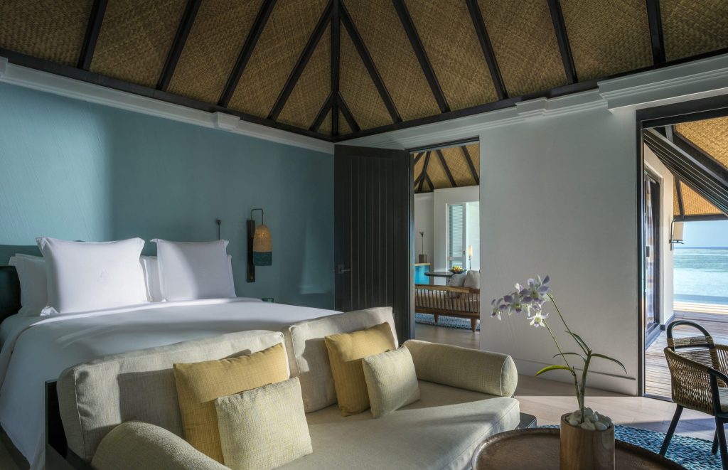 Hotelzimmer mit großem Bett, blau gestrichenen Wänden, Sofa, Strohdach und Meerblick durch offene Tür