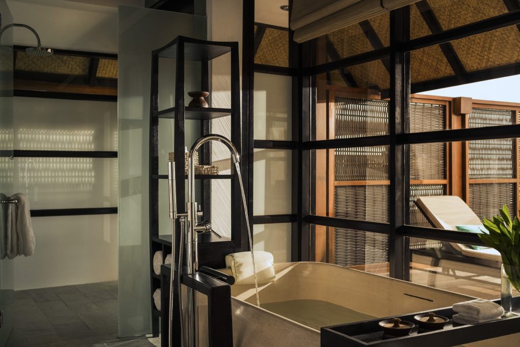 Badezimmer mit freistehender Badewanne aus hellem Stein, großem Fenster und Blick auf Liege im Außenbereich