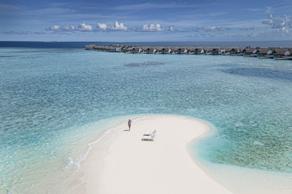 Sandbank im klaren türkisfarbenen Wasser mit zwei weißen Liegestühlen und einer Person, Bungalows im Hintergrund