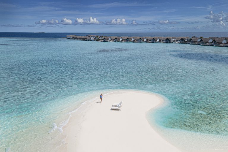 Sandbank im klaren türkisfarbenen Wasser mit zwei weißen Liegestühlen und einer Person, Bungalows im Hintergrund