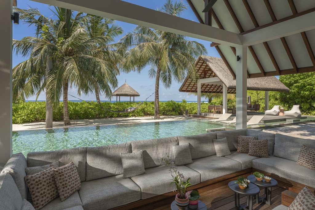 Außenlounge mit grauen Loungemöbeln und Pool vor Palmen im Four Seasons Resort Maldives at Landaa Giraavaru