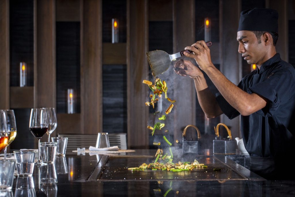 Koch bereitet Gemüse auf Teppanyaki-Platte zu vor Wand mit Holzelementen und warmer Beleuchtung im Four Seasons Resort.