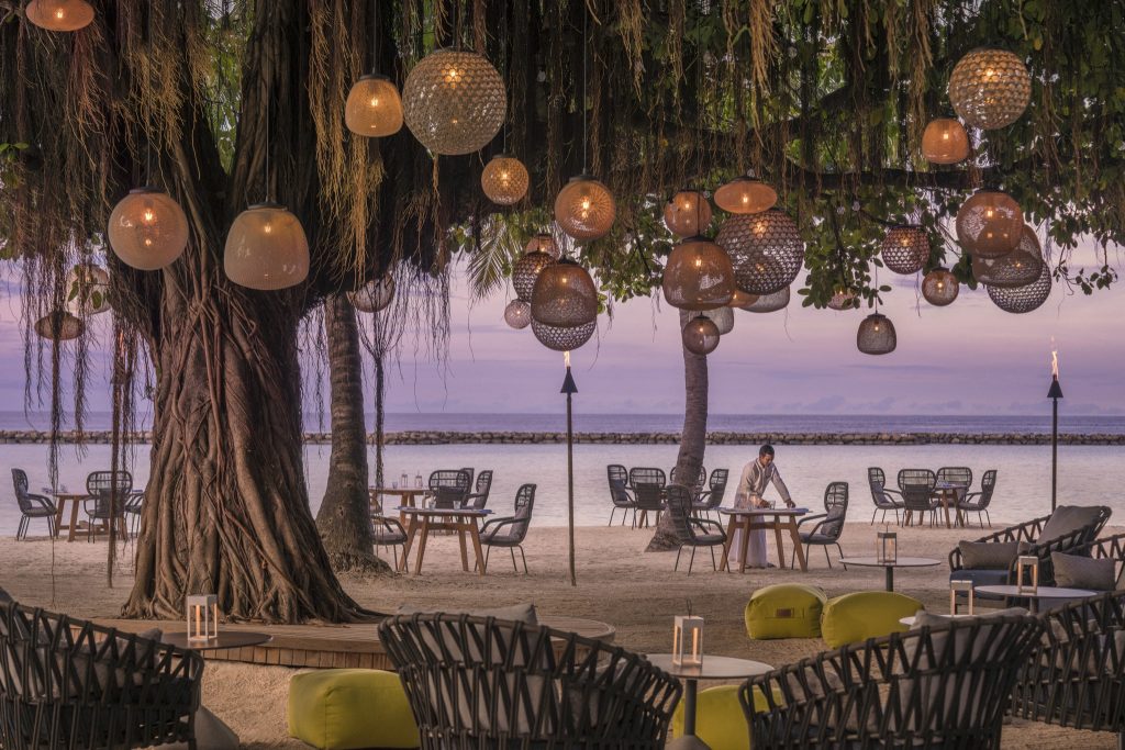Outdoor-Sitzlounge mit Rattanmöbeln und gelben Kissen unter Baum mit geflochtenen Lampen am Strand