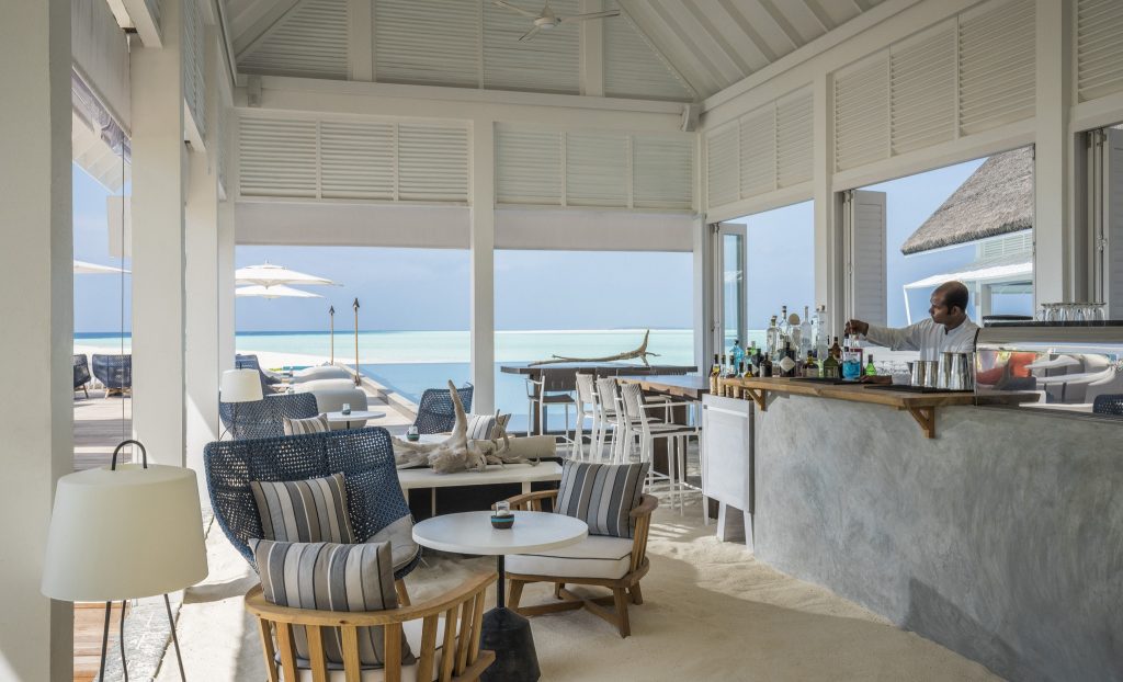Lichtdurchfluteter Lounge-Bereich mit hölzernen Möbeln, blauen Kissen, Bar mit Betonfläche und Blick auf Meer