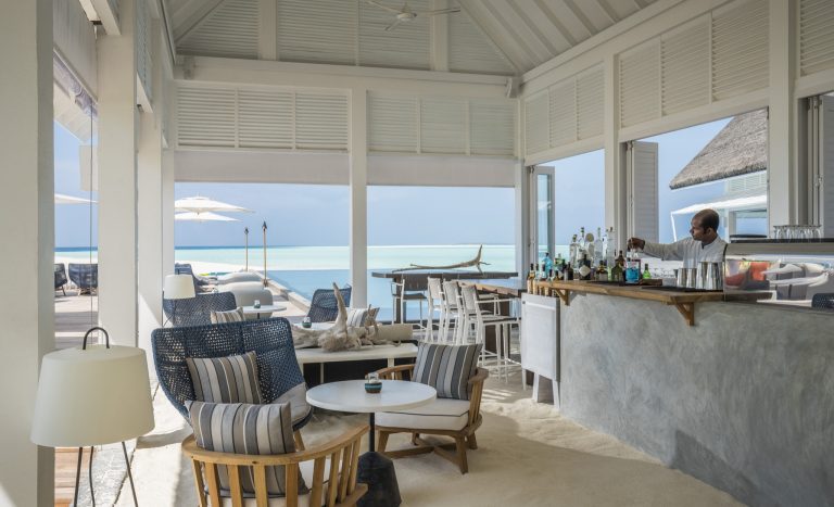 Lichtdurchfluteter Lounge-Bereich mit hölzernen Möbeln, blauen Kissen, Bar mit Betonfläche und Blick auf Meer
