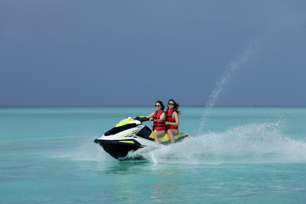 Zwei Personen in roten Rettungswesten auf weißem Wasserfahrzeug mit gelben Akzenten auf türkisblauem Meer unter bewölktem.