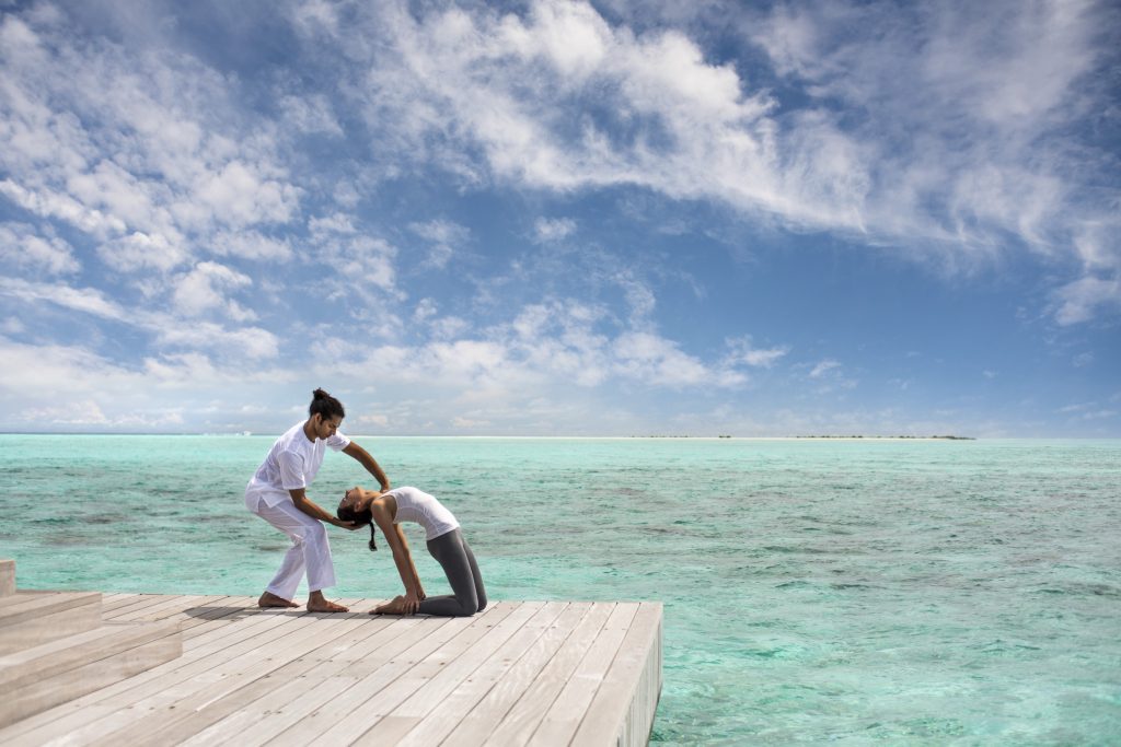 Zwei Personen in Yoga-Position auf hellem Holzsteg über türkisfarbenem Meer im Four Seasons Resort Maldives