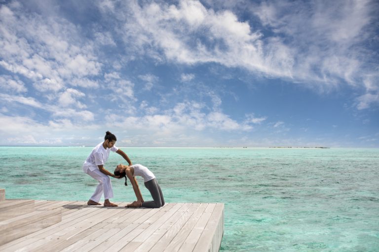 Zwei Personen in Yoga-Position auf hellem Holzsteg über türkisfarbenem Meer im Four Seasons Resort Maldives