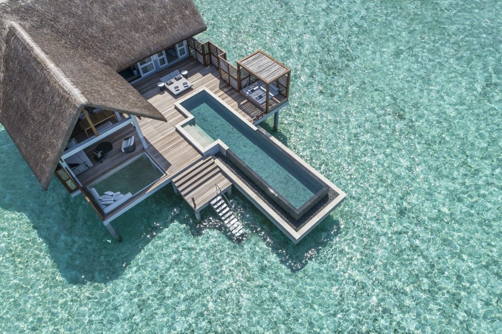 Überwasserbungalow mit Strohdach, Holzterrasse, langem Pool und weißen Liegestühlen über türkisfarbenem Meer
