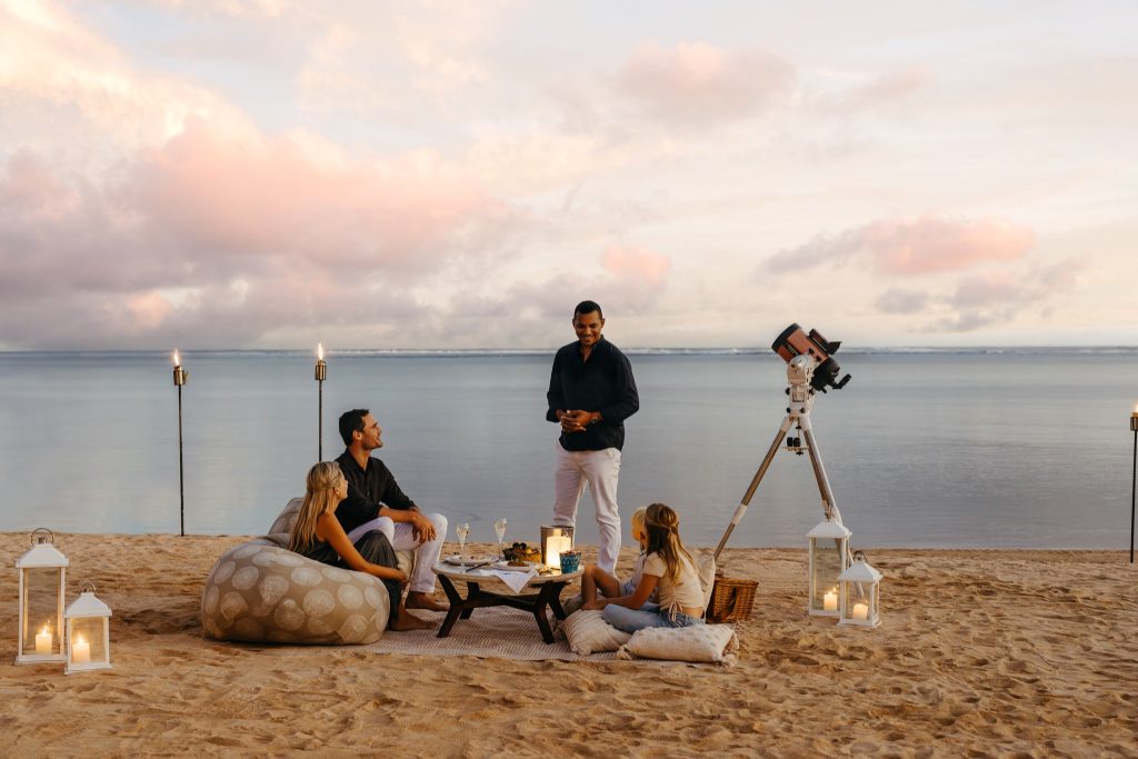 Gruppe von Menschen sitzt am Strandufer bei Heritage Le Telfair mit Kissen, Laterne, Fackeln und Teleskop am Abend