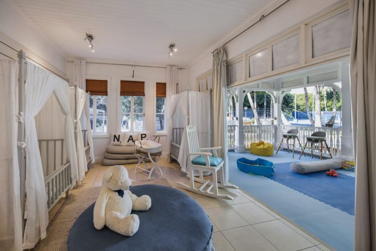 Kinderbereich mit zwei Babybetten, großem weißen Teddybären und Schaukelstuhl auf der Veranda im Heritage Le Telfair