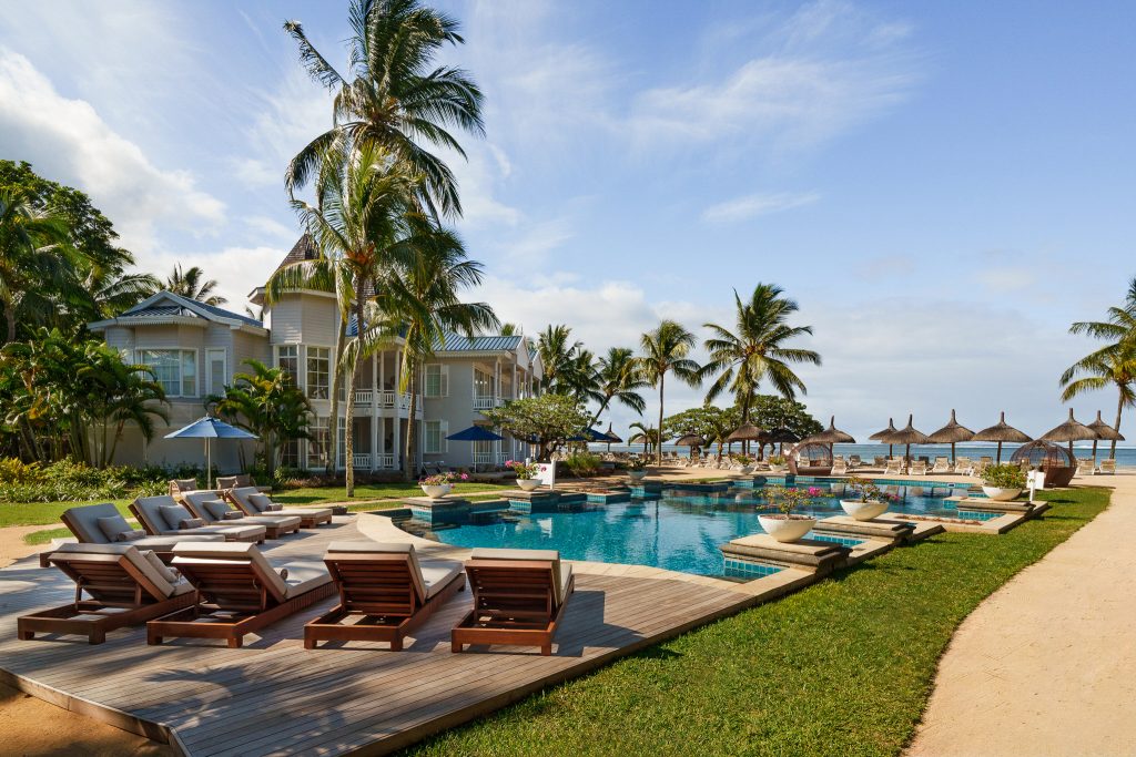 Poolbereich mit Holzdeck, beigen Liegestühlen, Palmen und Resort mit blauer Dachkonstruktion in Mauritius