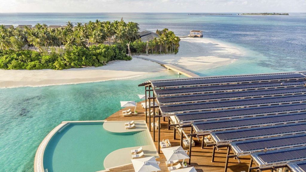 Poolbereich mit Holzdecks, Sonnenschirmen und Blick auf das türkisfarbene Meer mit Palmen im Hintergrund