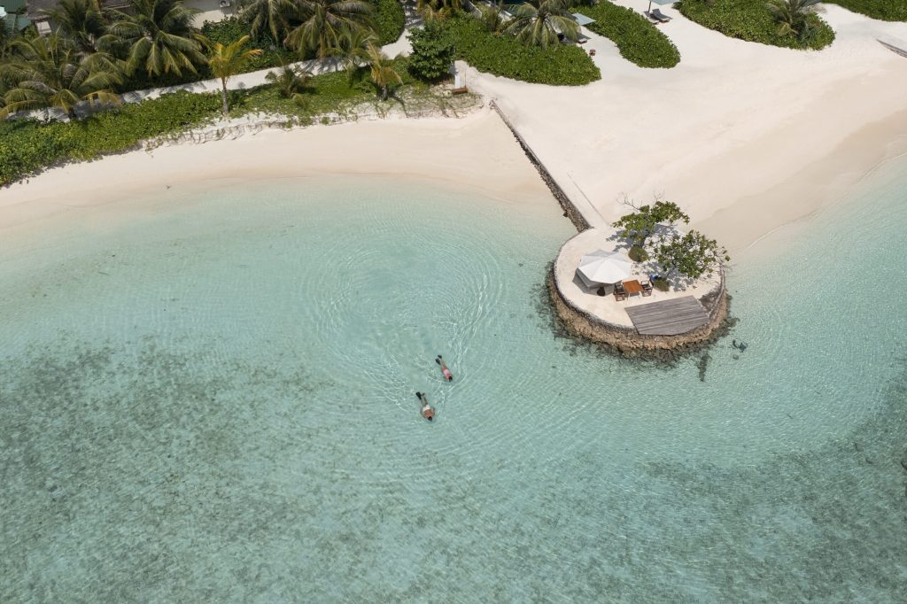 Luftaufnahme eines Strandes mit türkisfarbenem Wasser, zwei Personen im Meer bei runder Plattform mit Tisch und Sonnenschirm