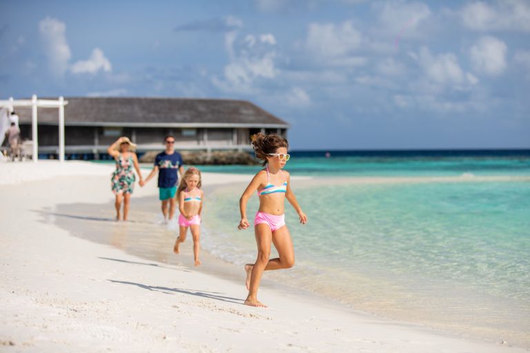 Familie mit Kindern spielt am weißen Sandstrand neben türkisblauem Meer bei Kagi Maldives Spa Island