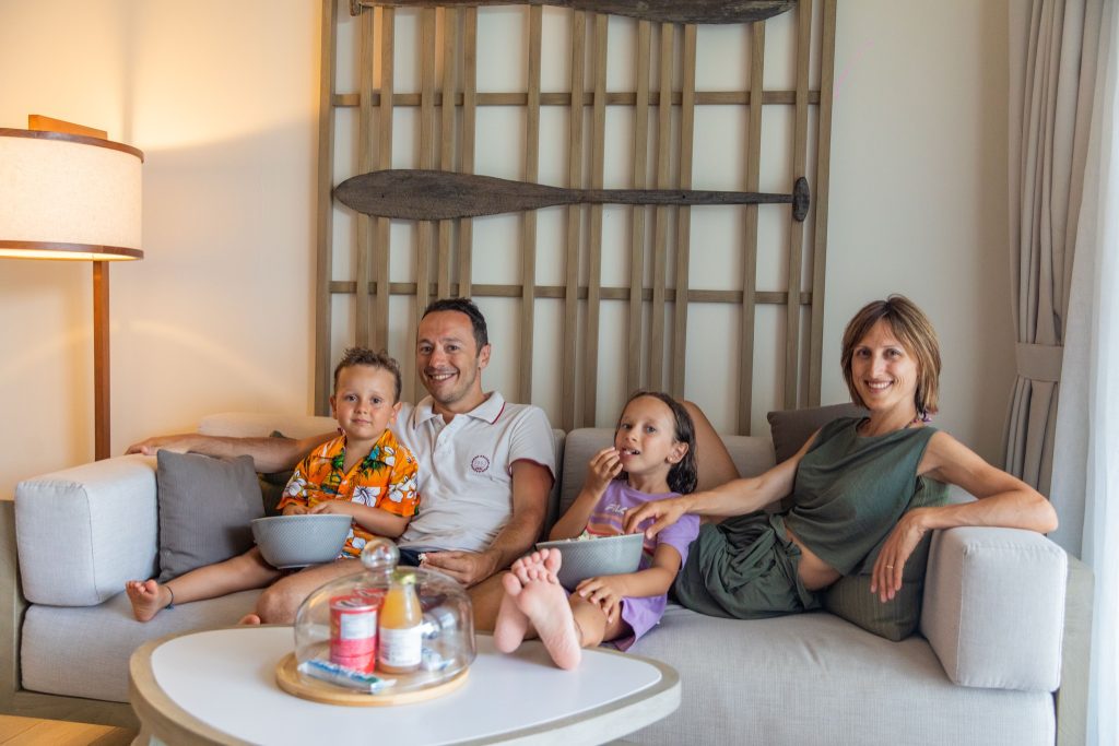 Wohnbereich mit Sofa, Holzstruktur und Paddeln an der Wand, Familie sitzt entspannt, Getränke auf Tisch