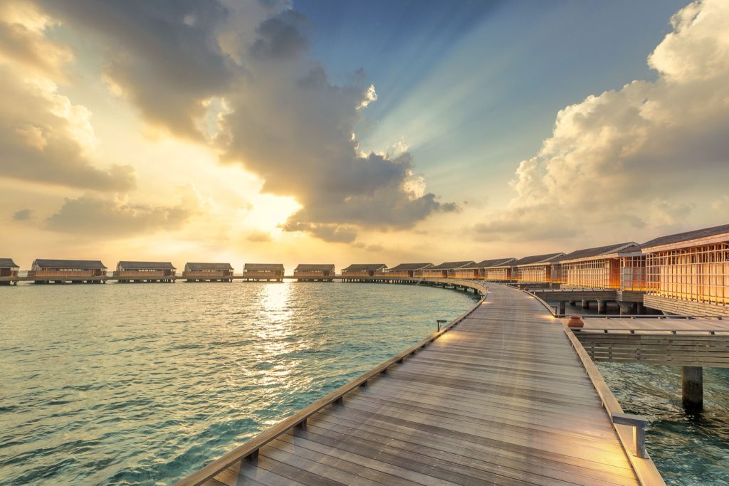 Reihe von Überwasser-Bungalows an einem geschwungenen Holzsteg bei Sonnenuntergang auf Kudadoo Private Island Malediven