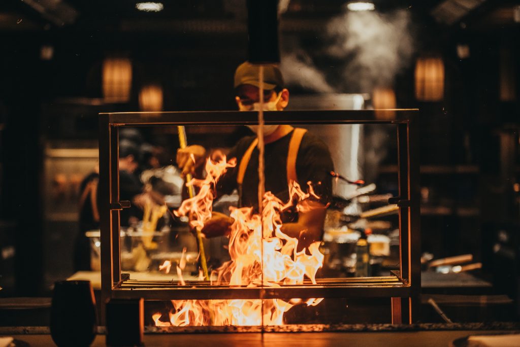 Offenes Kochszenario im Restaurant AI Kisu mit Feuerstelle und Koch im Hintergrund bei Dunkelbeleuchtung