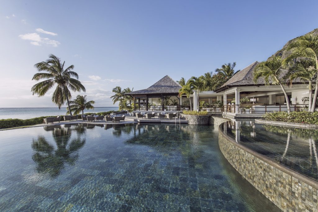 Großer klarer Pool mit blauen und grünen Fliesen, Palmen, Meer und traditionellen Pavillons im Hintergrund