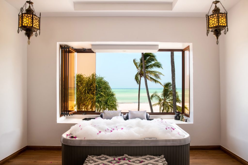 Hotelzimmer mit großem Whirlpool, Holzfußboden, zwei Metalllampen und Blick auf Meer und Palmen in Sansibar
