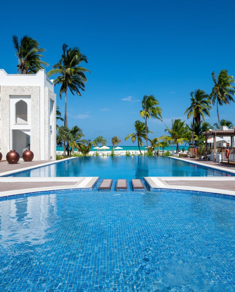 Poolbereich mit blau gefliesten Boden, weißem Steinbogen, Palmen und Sonnenliegen im Hotel Lux Marijani Sansibar