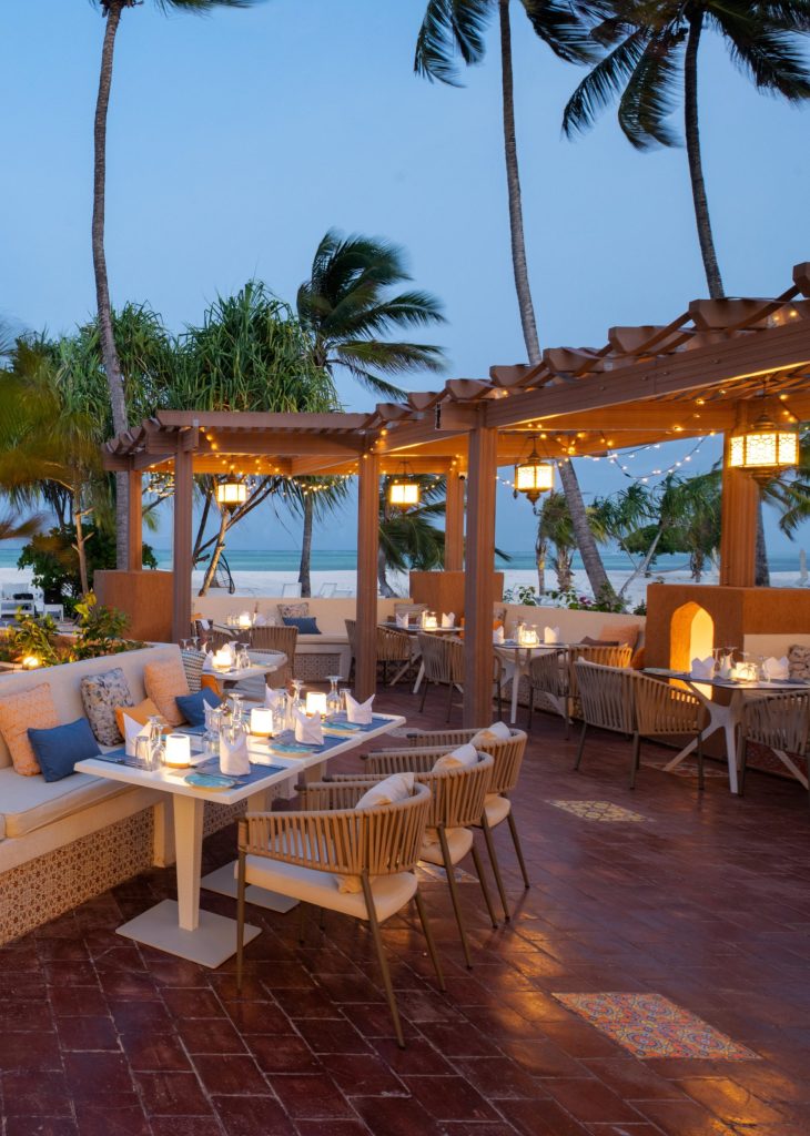 Restaurantterrasse mit Holz- und Kunstfasertischen, weißen Tischdecken, Laternen, Pergola mit Lichterketten und Palmen.