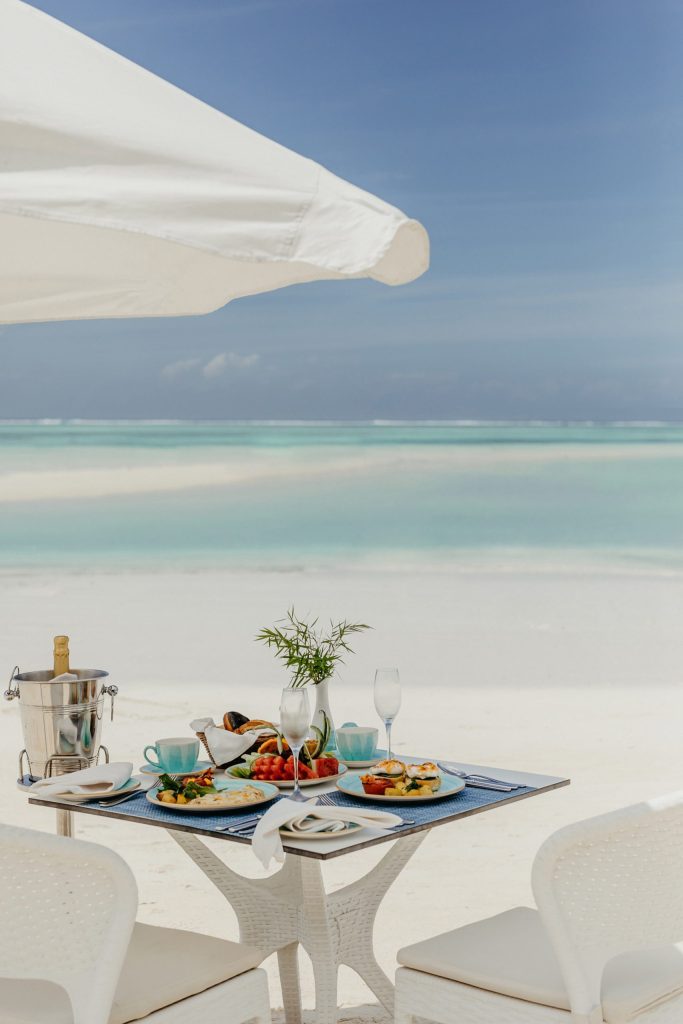 Gedeckter Tisch aus hellem Rattan mit Porzellantellern und Champagnerkühler am Sandstrand