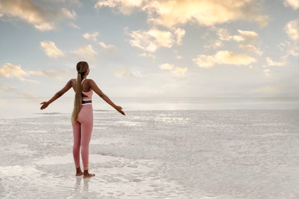 Person am Strand von hinten mit ausgestreckten Armen, nasser Sand und bewölkter Himmel bei Sonnenaufgang