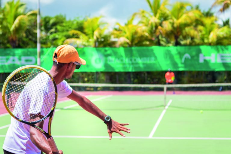 Tennisplatz mit roter und grüner Spielfläche umgeben von Palmen und Tennisspieler mit oranger Kappe
