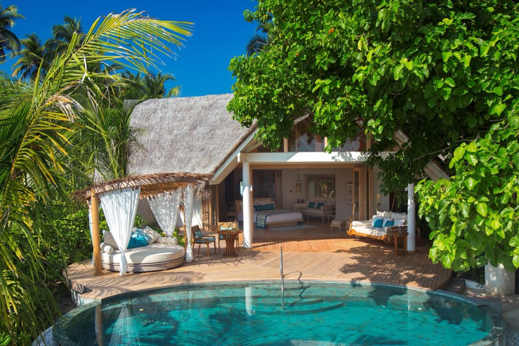Bungalow mit Strohdach, Holzterrasse, rundem Pool, Sitzecke unter Pavillon, helles Holzmobiliar und großes Bett