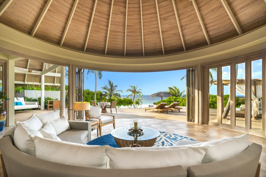 Innenraum der Beach Residence mit gebogenem cremeweißem Sofa, Glastisch und Blick auf Terrasse und Meer