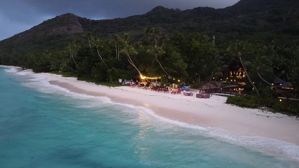 Strand mit weißem Sand, türkisfarbenem Meer, Holztischen mit Lichterketten und Palmen am Rand