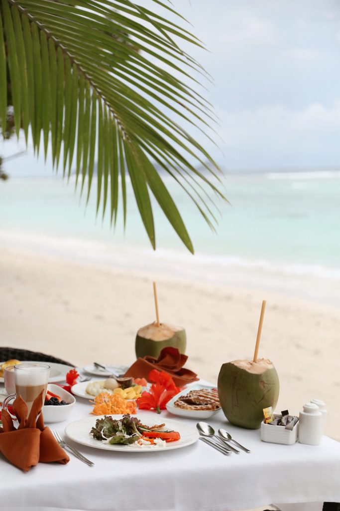 Gedeckter Tisch am Strand mit Salat, Spiegeleiern, Obst, Kokosnüssen und Besteck vor Sandstrand und Palme