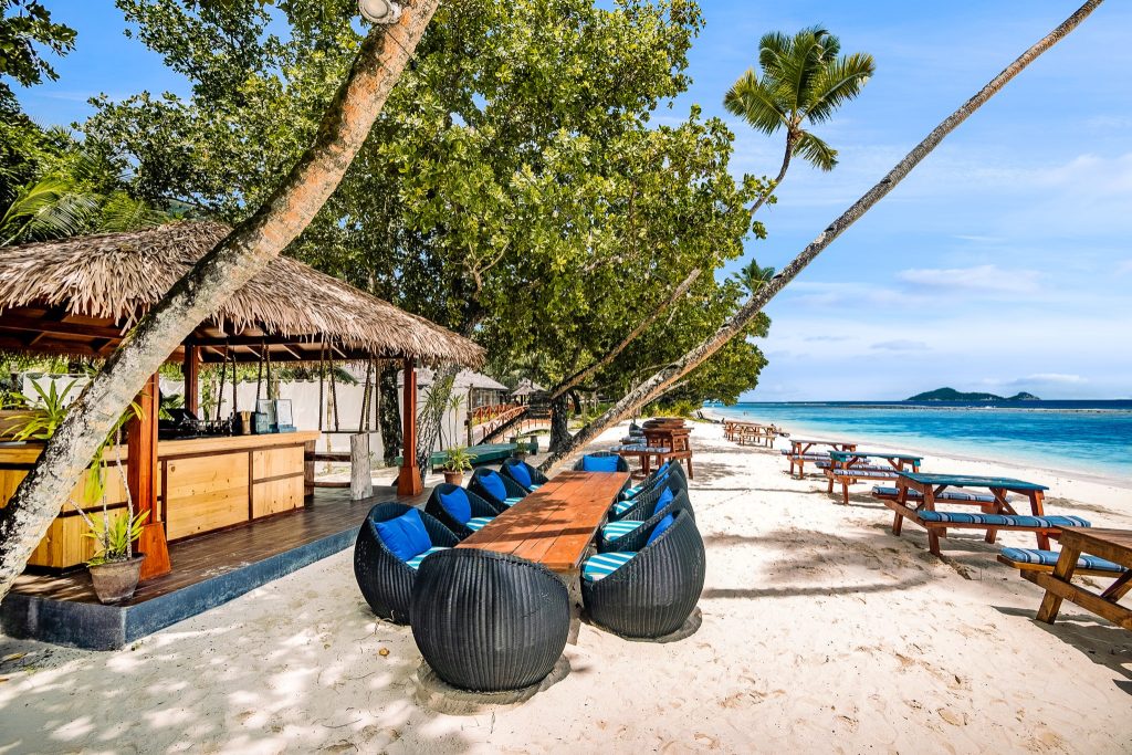 Strandbar am Sandstrand mit Holztisch, blauen Kissen auf geflochtenen Sesseln und Palmstrohdach in hellem Tageslicht