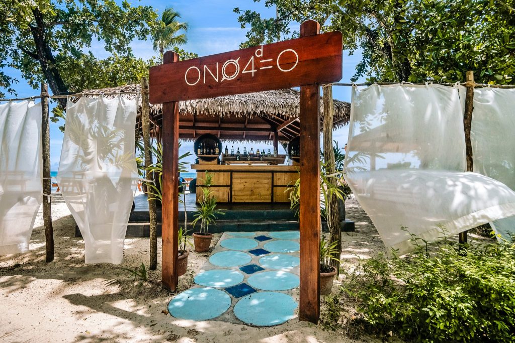 Outdoor-Bar mit Strohdach und Holzeingang, umgeben von Pflanzen und weißen Vorhängen bei Tageslicht