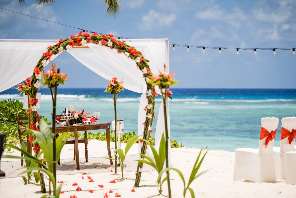 Strandbereich mit weißem Baldachin, roter und weißer Blumendekoration, Tisch und Stühlen mit roten Schleifen am Meer