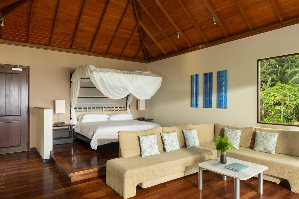 Zimmer mit cremefarbenem Ecksofa, blau gemusterten Kissen, Baldachinbett auf Plattform und Blick auf Dschungellandschaft