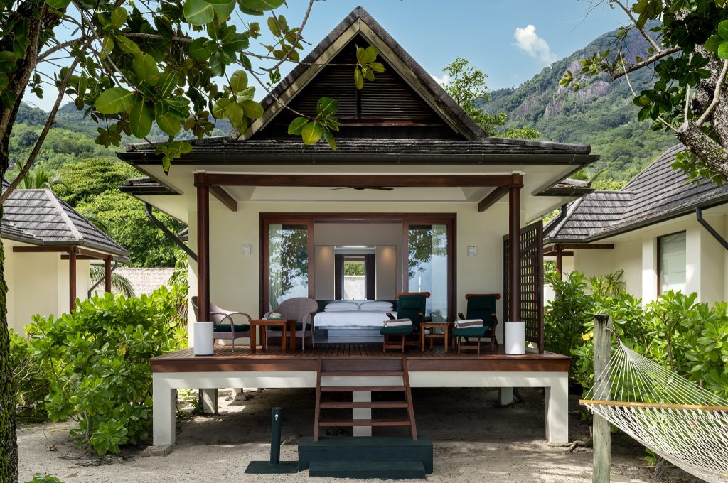 Tropische Beach Villa mit Holzveranda, Holzstühlen, Glasfront und Hängematte umgeben von Grün und Bergen auf den