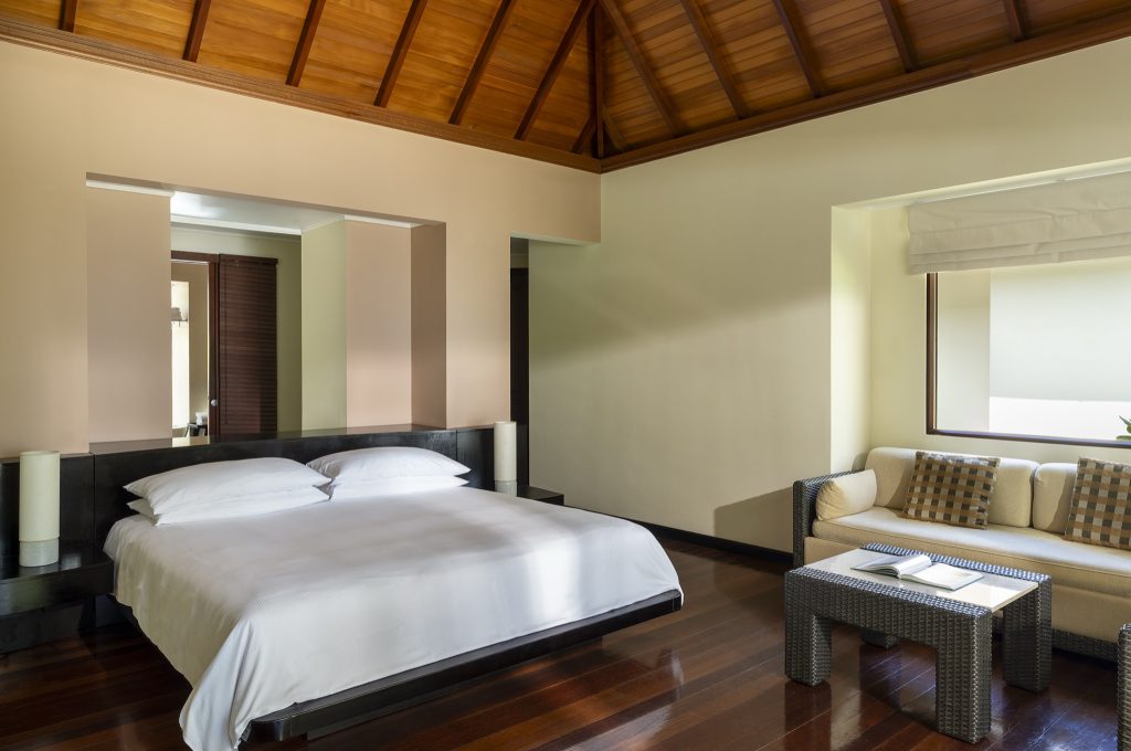 Geräumiges Beach Villa Zimmer mit Doppelbett auf Holzpodest, Couch, geflochtenem Tisch und Holzdielenboden