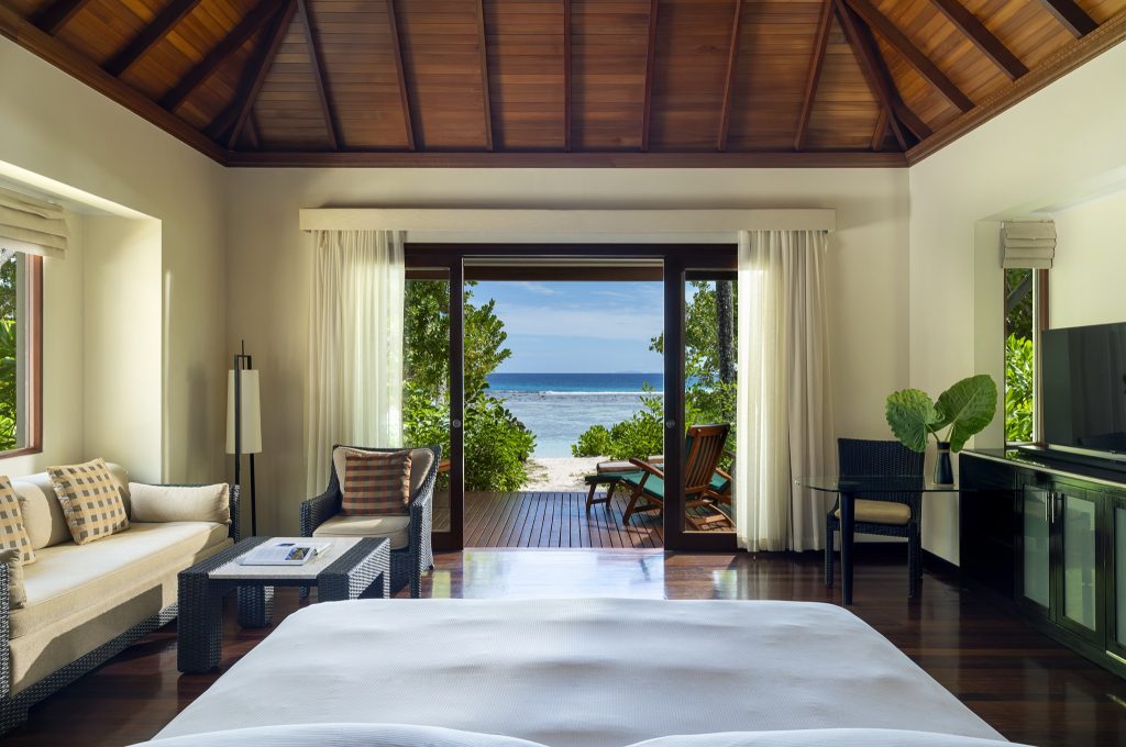 Zimmer der Beach Villa mit weißem Bett, Rattan-Sitzecke, dunklem Holzboden und Meerblick auf den