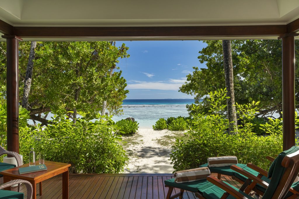 Terrasse der Beach Villa im NIVA Labriz Seychelles mit zwei Holzliegen, Holztisch, grünem Pflanzenrahmen und Blick.